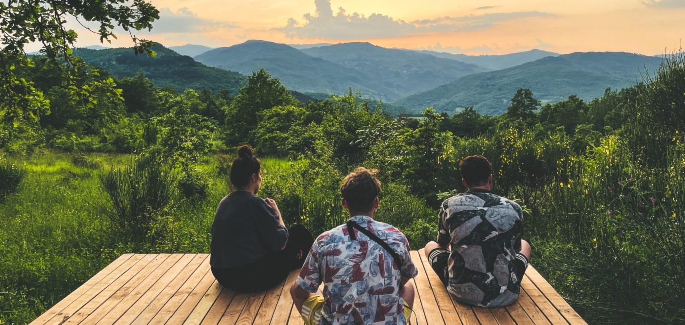 Drei Personen blicken auf eine hügelige Landschaft bei Sonnenuntergang.